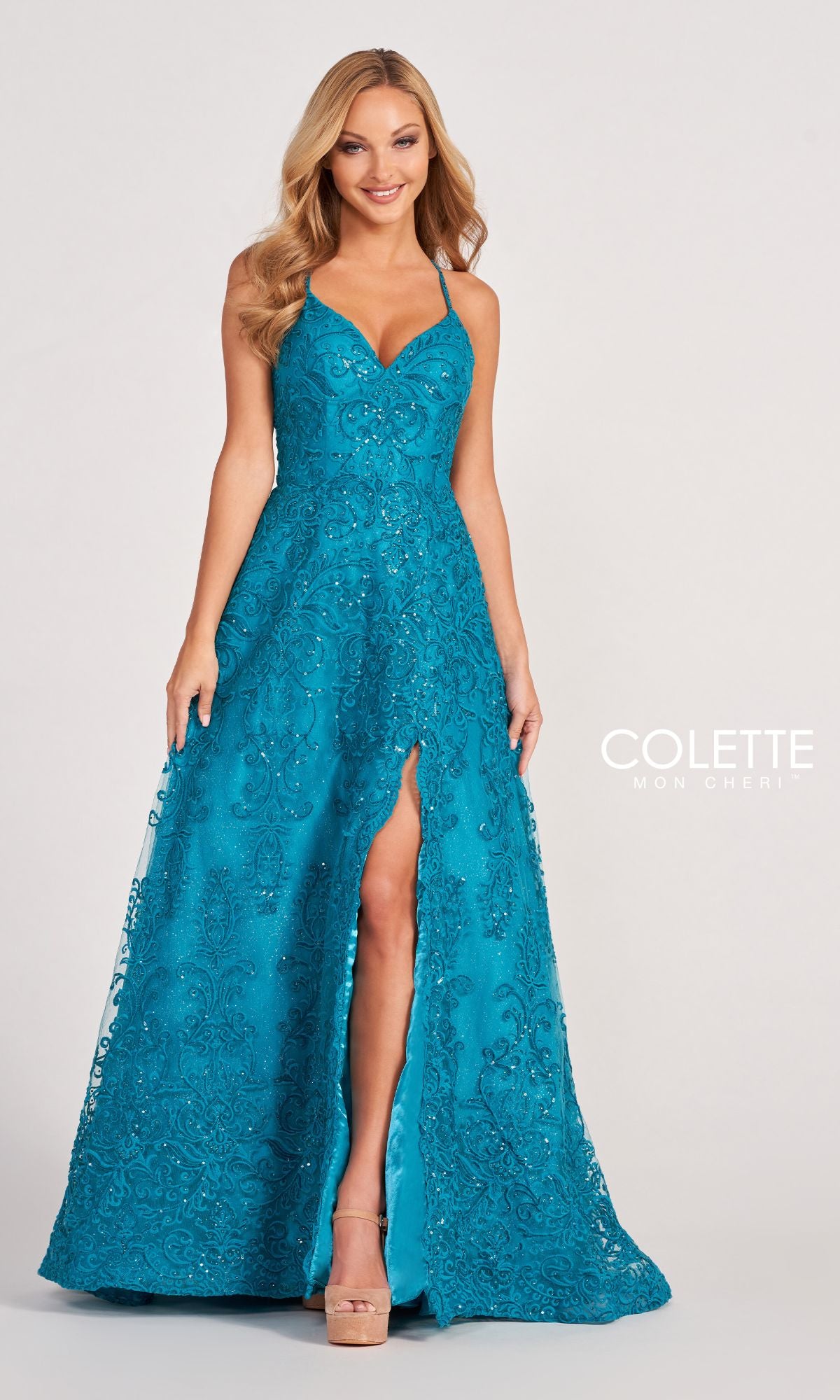 A woman faces the camera in a teal, beaded Colette CL2028 Strappy-Back Lace A-Line Prom Dress featuring thin straps, a high slit, and intricate embellishments. The gown has an open back and is ideal for prom or evening events.